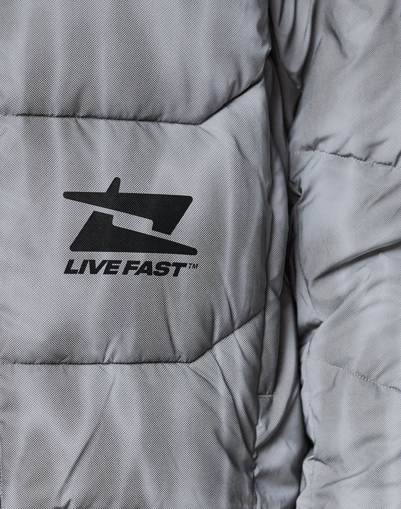 Close-up of the Live Fast Puffer Jacket by LIVE FAST DIE YOUNG, a gray, water-repellent quilted jacket with a black airplane-inspired logo above "LIVE FAST™" printed on the fabric.