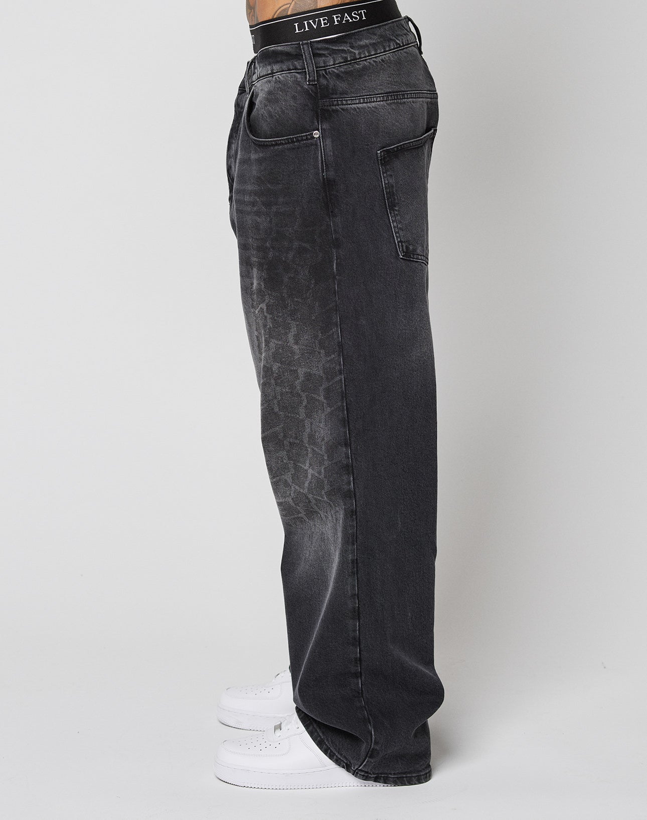 A person in LFDY Tire Tracks Denim by LIVE FAST DIE YOUNG, featuring loose-fit faded black jeans with subtle tire track patterns and a black LIVE FAST waistband, stands sideways against a plain light background in white sneakers.