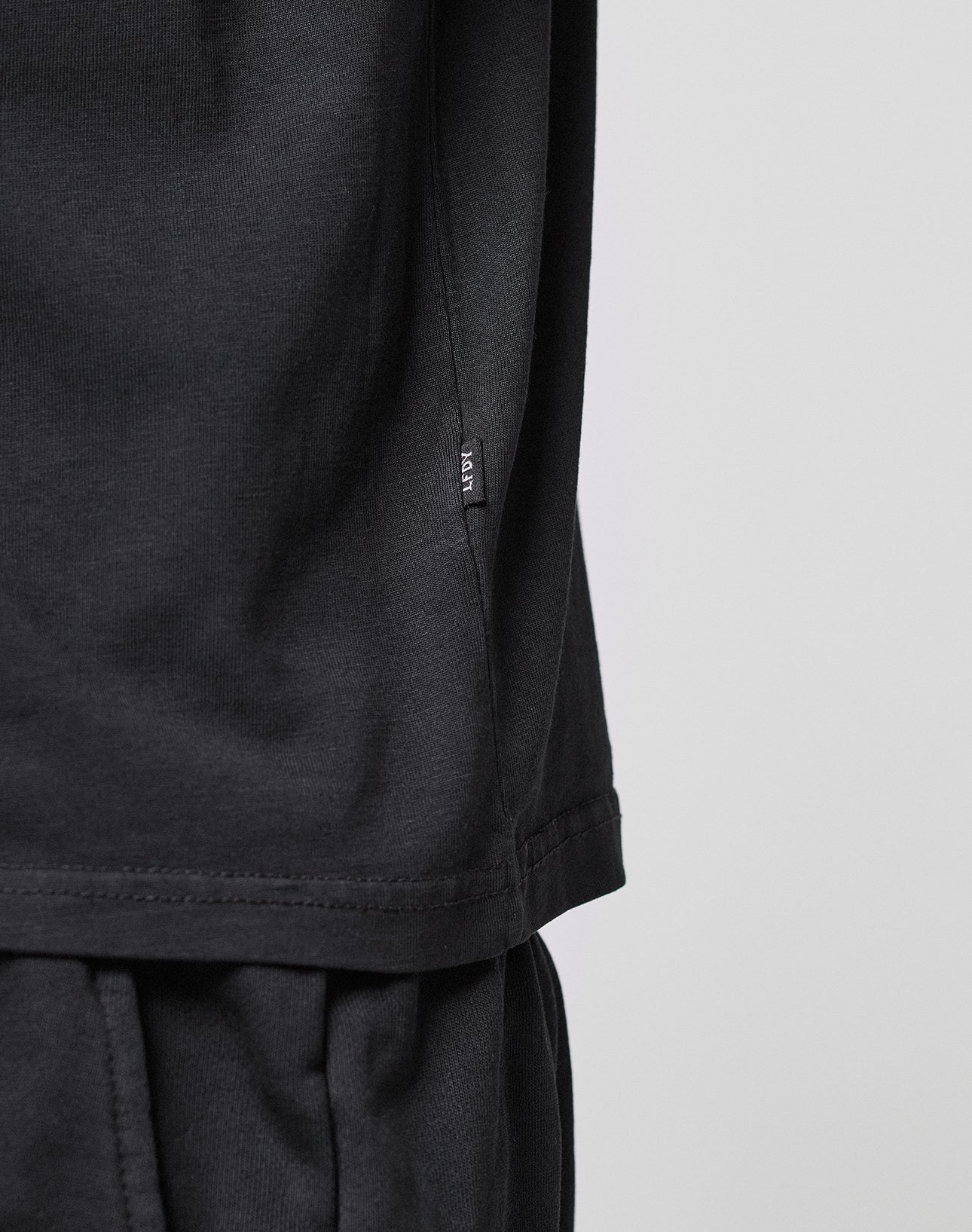 Close-up of a person wearing the LIVE FAST DIE YOUNG Basic LiveFast Tee in black and black pants, featuring a small black tag near the shirt’s side seam against a plain, light background.
