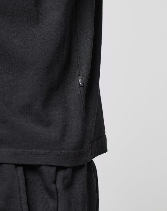 Close-up of a person wearing the LIVE FAST DIE YOUNG Basic LiveFast Tee in black and black pants, featuring a small black tag near the shirt’s side seam against a plain, light background.