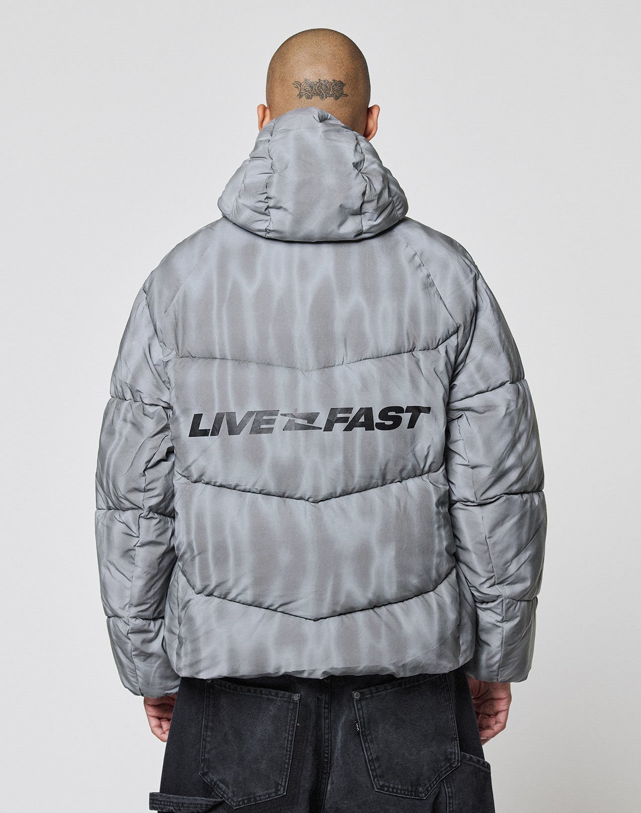 A person with a shaved head, seen from behind, wears the LIVE FAST DIE YOUNG Live Fast Puffer Jacket—a shiny gray, water-repellent, oversized reflective jacket with a hood and "LIVE FAST" in bold black letters on the back.