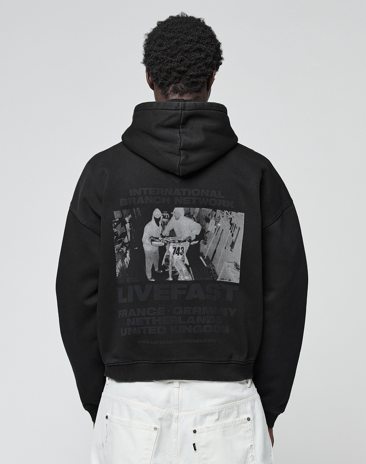 A person wears the LFDY Grand Theft Hooded Zip by LIVE FAST DIE YOUNG, an oversize fit black hoodie with Kapuze, grayscale runner photo, “LIVE FAST” text, and country names. Made from soft Baumwolle for all-day comfort. | model