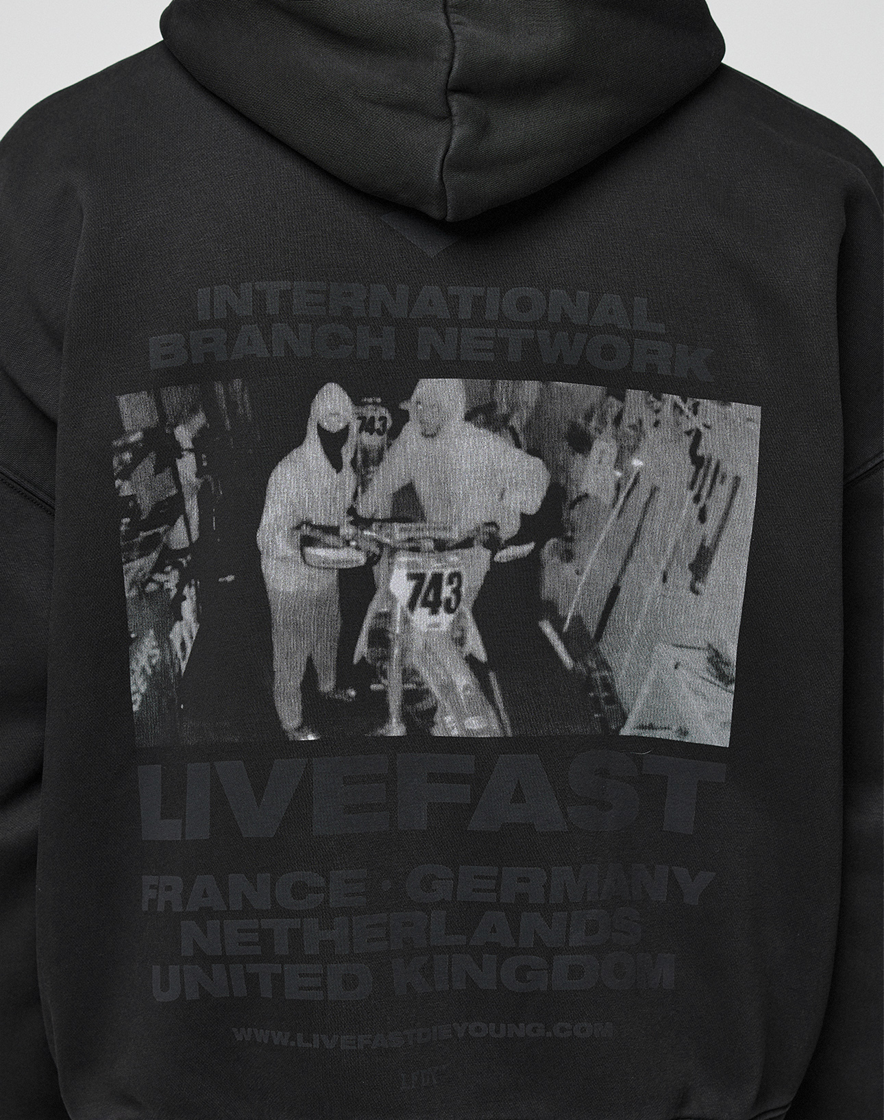 The LFDY Grand Theft Hooded Zip by LIVE FAST DIE YOUNG is an oversized black cotton hoodie with a grayscale print of two people by a motorbike (No. 743) and text: INTERNATIONAL BRANCH NETWORK LIVEFAST FRANCE · GERMANY NETHERLANDS UNITED KINGDOM.