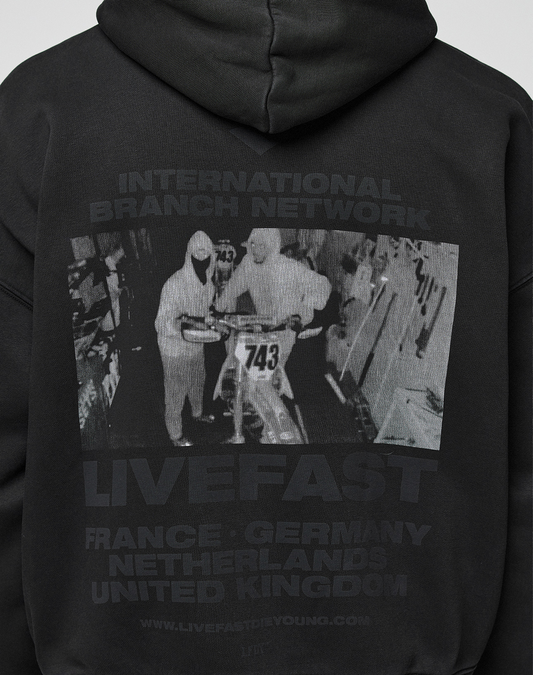 The LFDY Grand Theft Hooded Zip by LIVE FAST DIE YOUNG is an oversized black cotton hoodie with a grayscale print of two people by a motorbike (No. 743) and text: INTERNATIONAL BRANCH NETWORK LIVEFAST FRANCE · GERMANY NETHERLANDS UNITED KINGDOM.