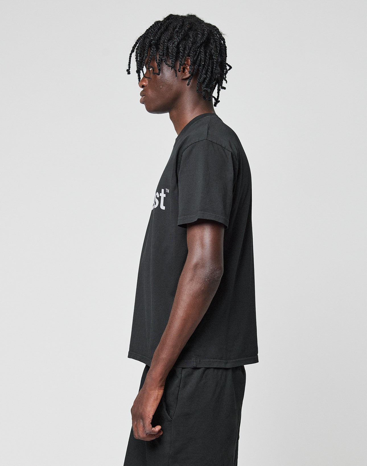 A person with short, twisted hair wears the LIVE FAST DIE YOUNG Basic LiveFast Tee in black and matching shorts, standing facing left against a plain light gray background.