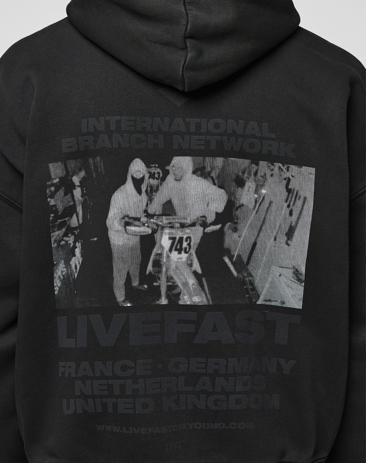 The LFDY Grand Theft Hooded Zip by LIVE FAST DIE YOUNG is an oversized black cotton hoodie with a grayscale print of two people by a motorbike (No. 743) and text: INTERNATIONAL BRANCH NETWORK LIVEFAST FRANCE · GERMANY NETHERLANDS UNITED KINGDOM.