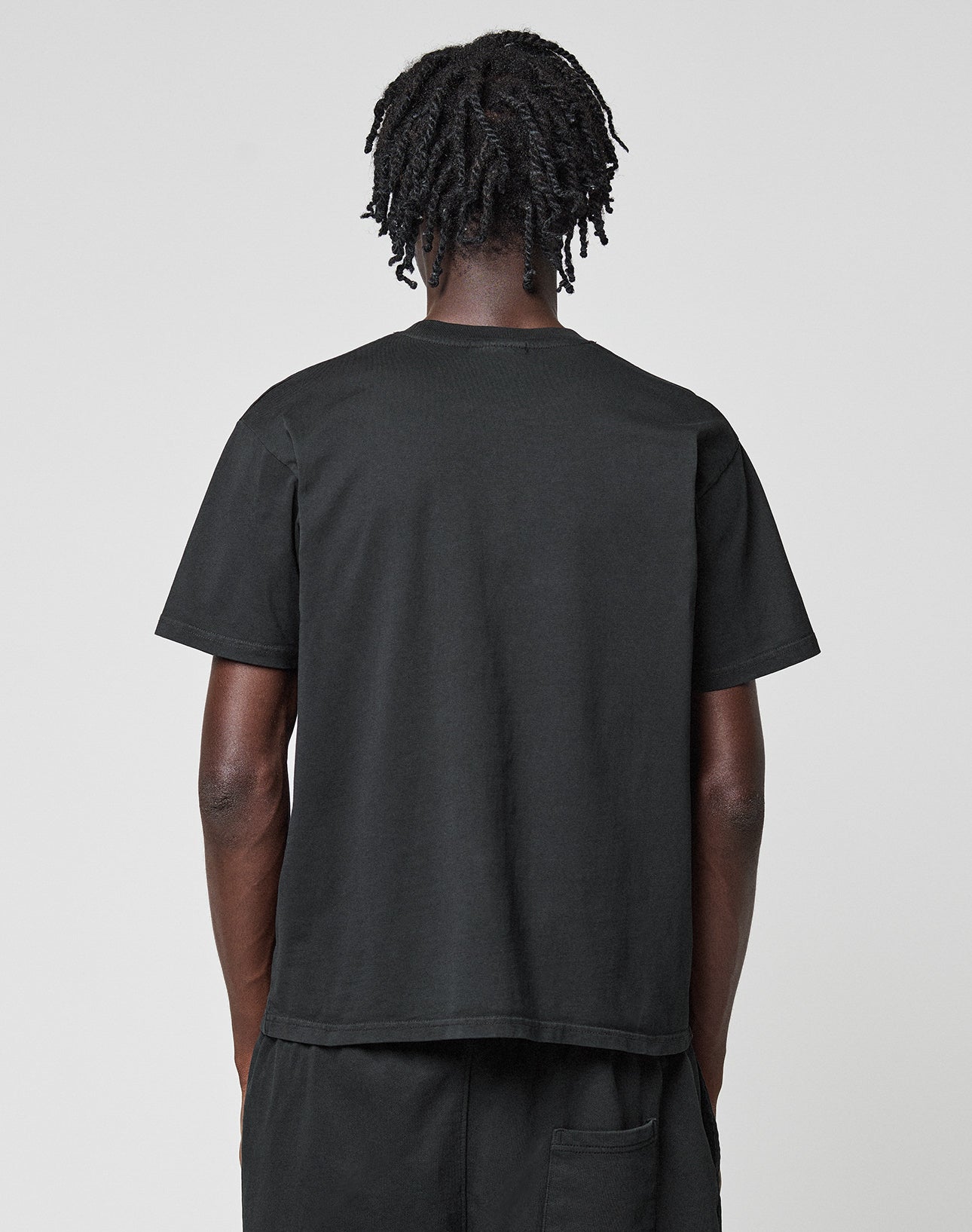 A person with short, twisted hair stands with their back to the camera, wearing a plain black Basic LiveFast Tee by LIVE FAST DIE YOUNG and black pants against a light gray background.