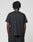 A person with short, twisted hair stands with their back to the camera, wearing a plain black Basic LiveFast Tee by LIVE FAST DIE YOUNG and black pants against a light gray background.