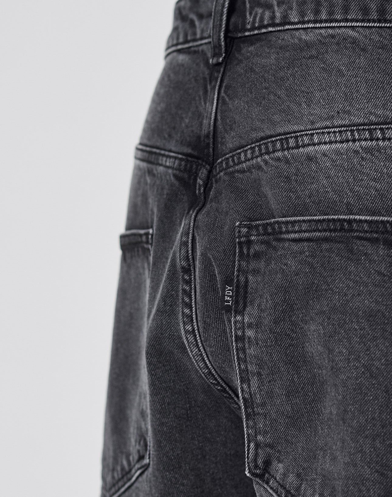 Close-up of the back of a person wearing LIVE FAST DIE YOUNG LFDY Tire Tracks Denim in a Baggy Fit, featuring faded black denim, visible seams, stitching, back pockets, and a small fabric tag on one pocket.