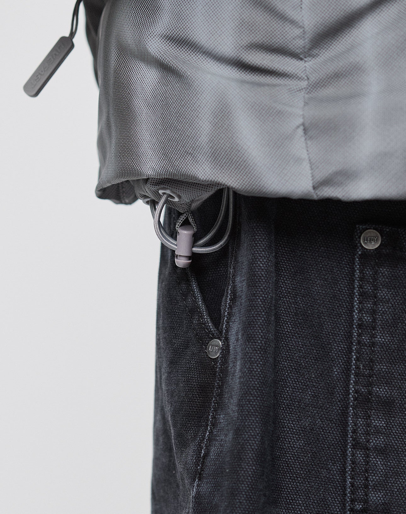 Close-up of the hem of the gray Live Fast Puffer Jacket by LIVE FAST DIE YOUNG, featuring an adjustable drawstring, visible toggle, and textured fabric, styled over dark pants.