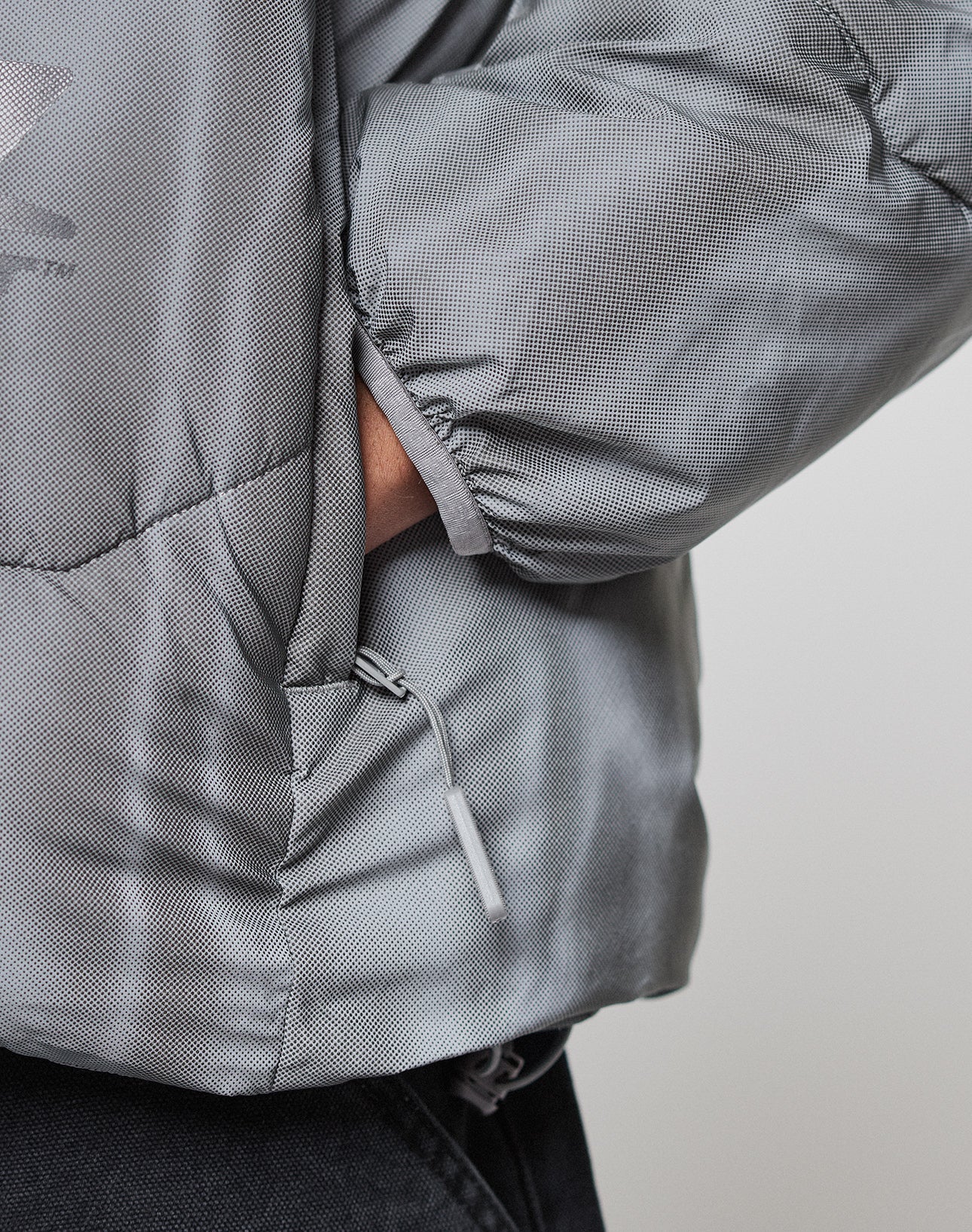 A close-up of a hand in the side pocket of the light gray, textured Live Fast Puffer Jacket by LIVE FAST DIE YOUNG, featuring a visible zipper and elastic cuff, paired with dark pants.