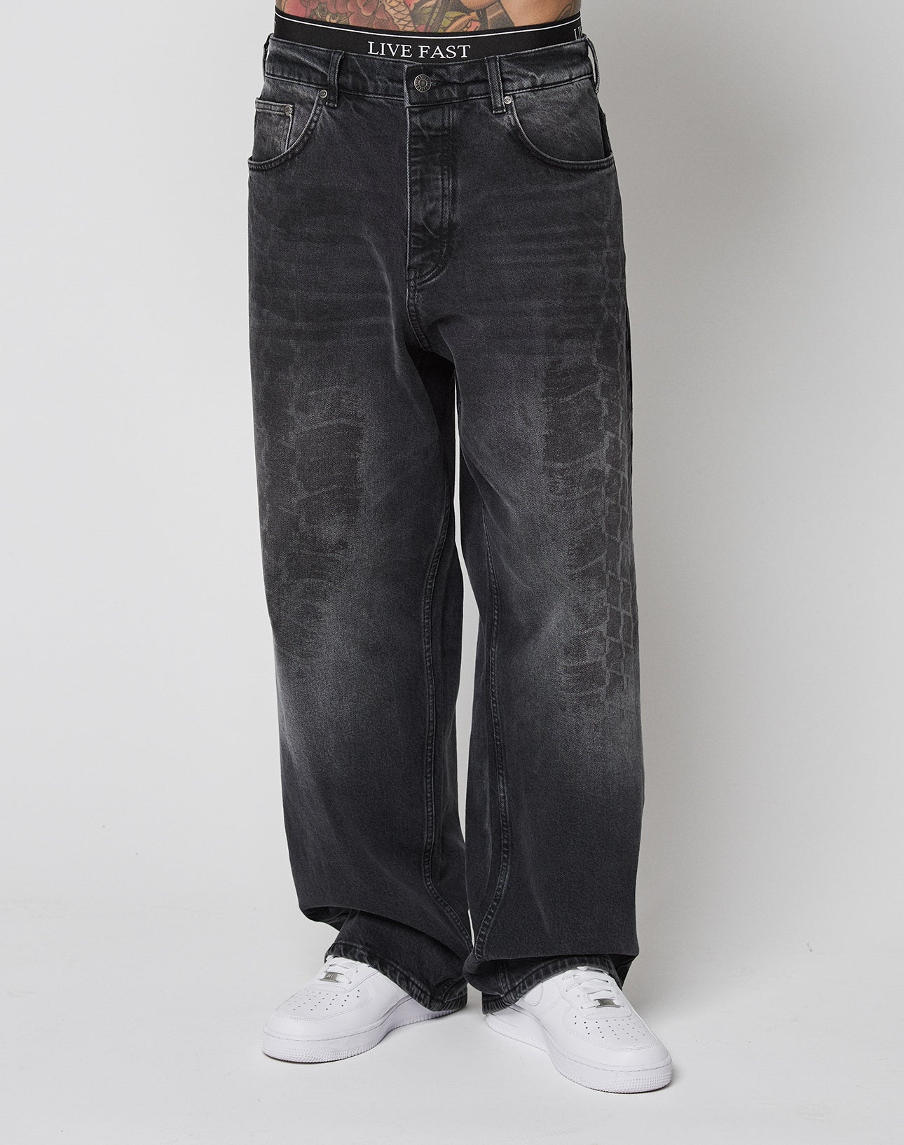 A person wearing LIVE FAST DIE YOUNG LFDY Tire Tracks Denim in a baggy fit with a visible LIVE FAST waistband and white sneakers, shown from the waist down against a plain white background. Made from 100% Baumwolle.