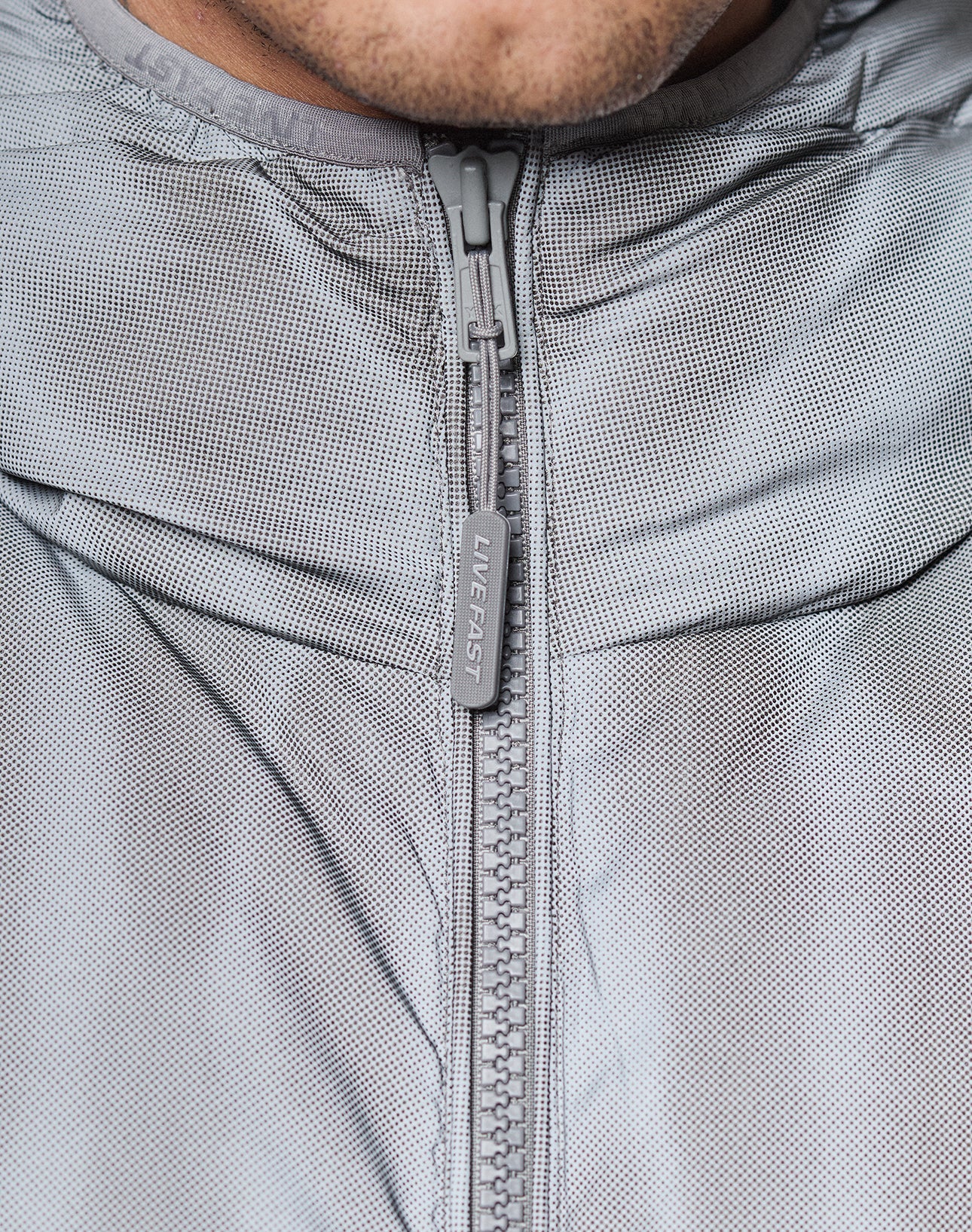Close-up of the Live Fast Puffer Jacket by LIVE FAST DIE YOUNG in gray, featuring water-repellent fabric, a large zipper with "LIVFAST" on the pull, and part of a person’s lower face visible at the top.