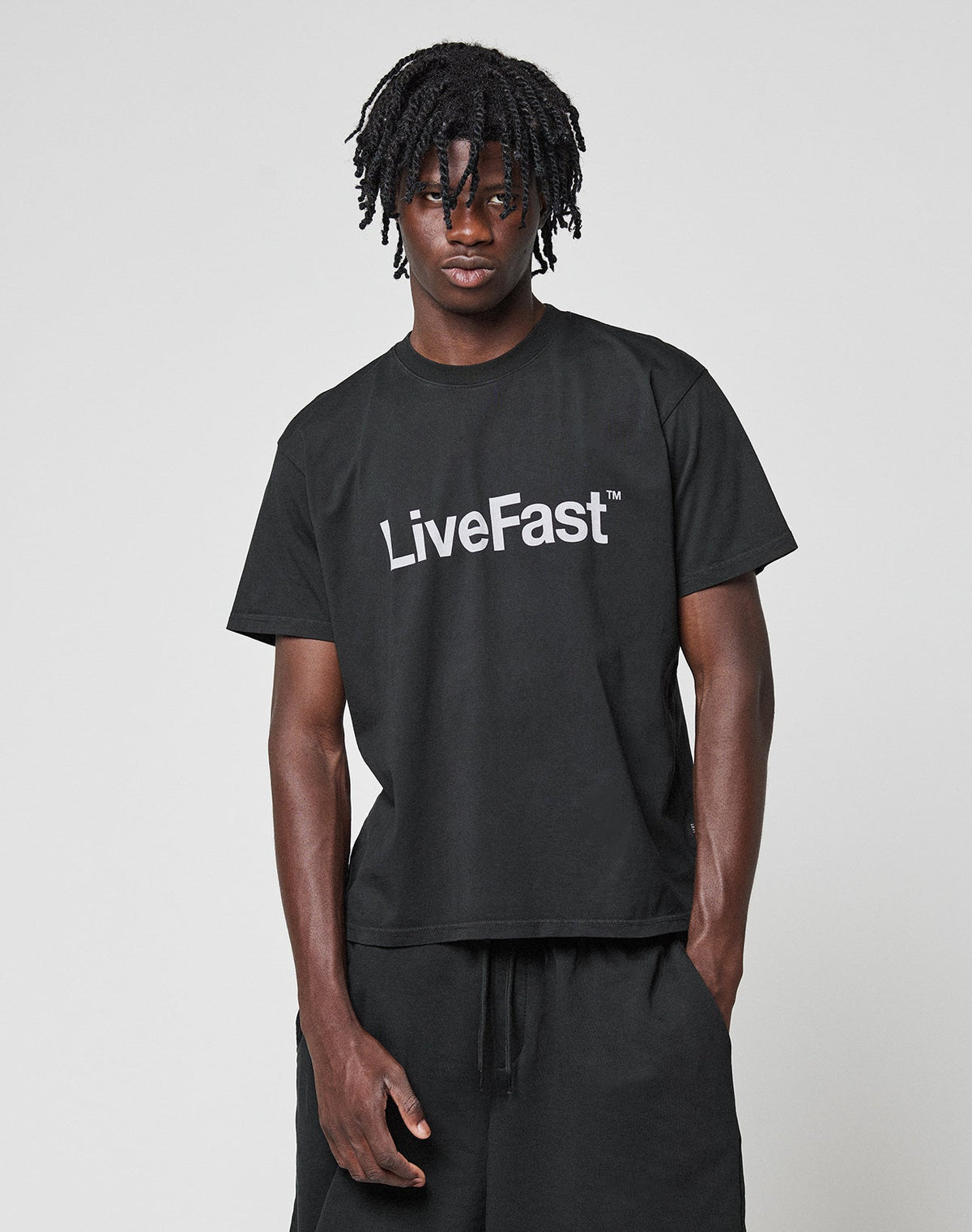 A person stands against a plain background wearing the LIVE FAST DIE YOUNG Basic LiveFast Tee in black featuring a bold LiveFast front print, paired with matching black pants.