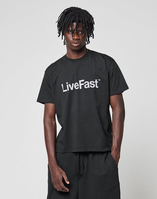 A person stands against a plain background wearing the LIVE FAST DIE YOUNG Basic LiveFast Tee in black featuring a bold LiveFast front print, paired with matching black pants.