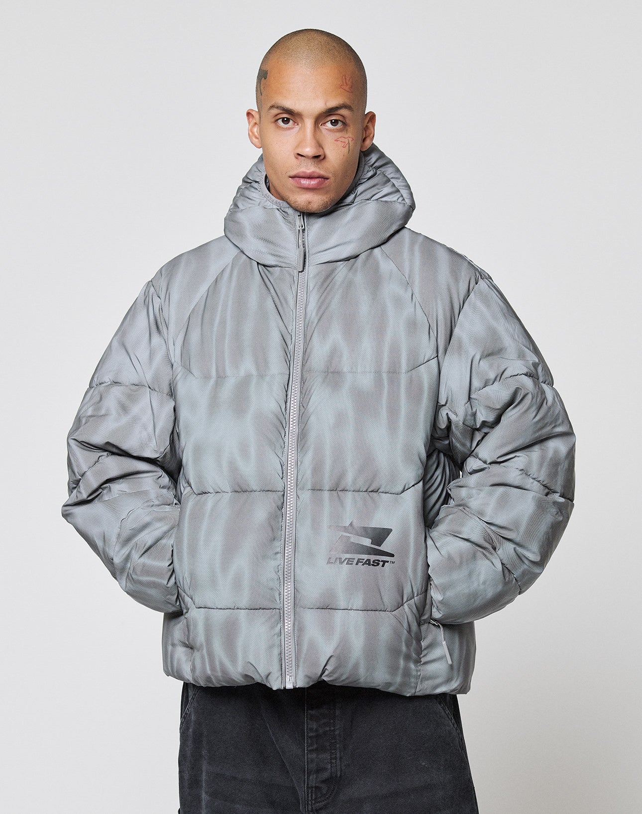 A person in a shiny, light grey Live Fast Puffer Jacket by LIVE FAST DIE YOUNG with high collar and pockets stands against a white background. The jacket features a subtle pattern, LIVE FAST logo, and is wasserabweisend for protection.