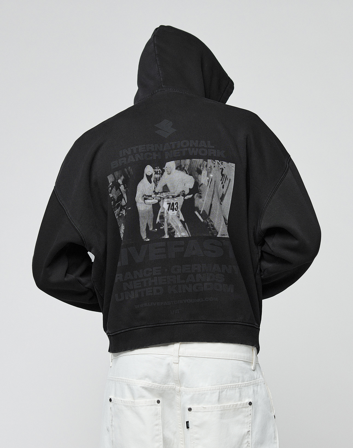 Someone faces away from the camera in a black, oversized LIVE FAST DIE YOUNG LFDY Grand Theft Hooded Zip aus Baumwolle with a grayscale back graphic of three people (one labeled 743) and country names, paired with white pants.