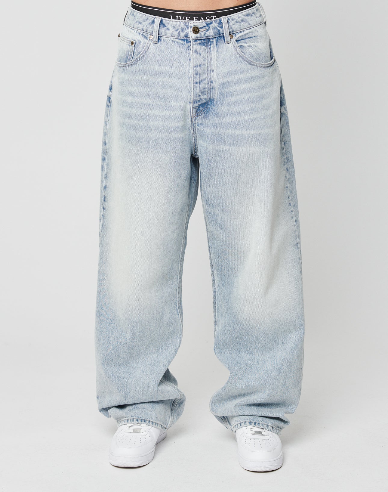 A person wears LFDY Baggy Denim by LIVE FAST DIE YOUNG—loose, light-wash blue jeans with a wide straight leg in 100% cotton. Only the lower torso and legs are shown, paired with white sneakers against a plain white background.