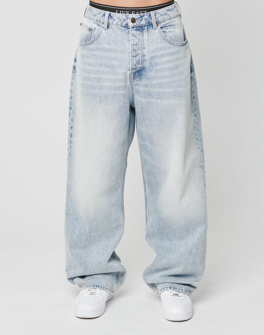 A person wears LFDY Baggy Denim by LIVE FAST DIE YOUNG—loose, light-wash blue jeans with a wide straight leg in 100% cotton. Only the lower torso and legs are shown, paired with white sneakers against a plain white background.