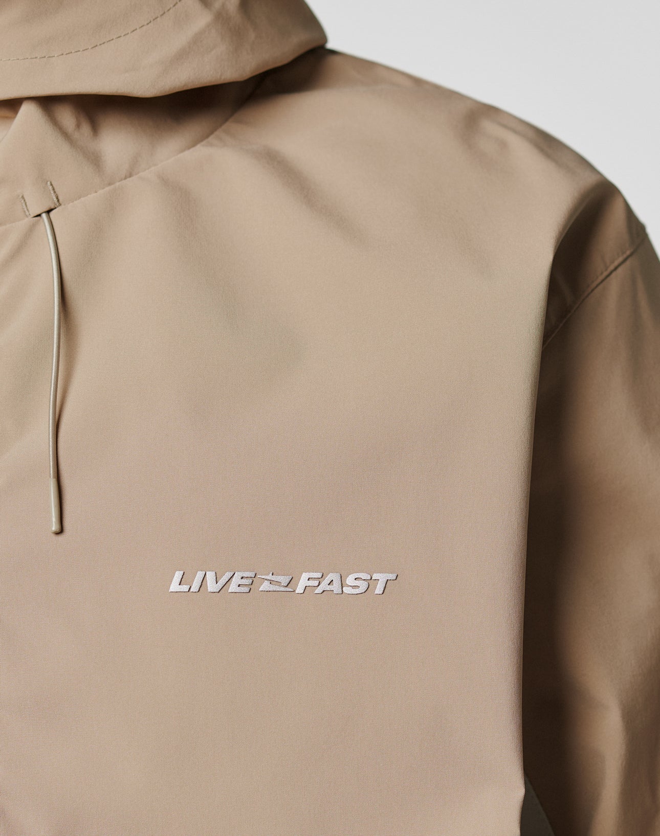 A close-up of the beige Basic Shell Jacket by LIVE FAST DIE YOUNG, featuring LIVE FAST and a race flag embroidered in white on the chest, with an oversize fit and a water-repellent zipper for added style and function.