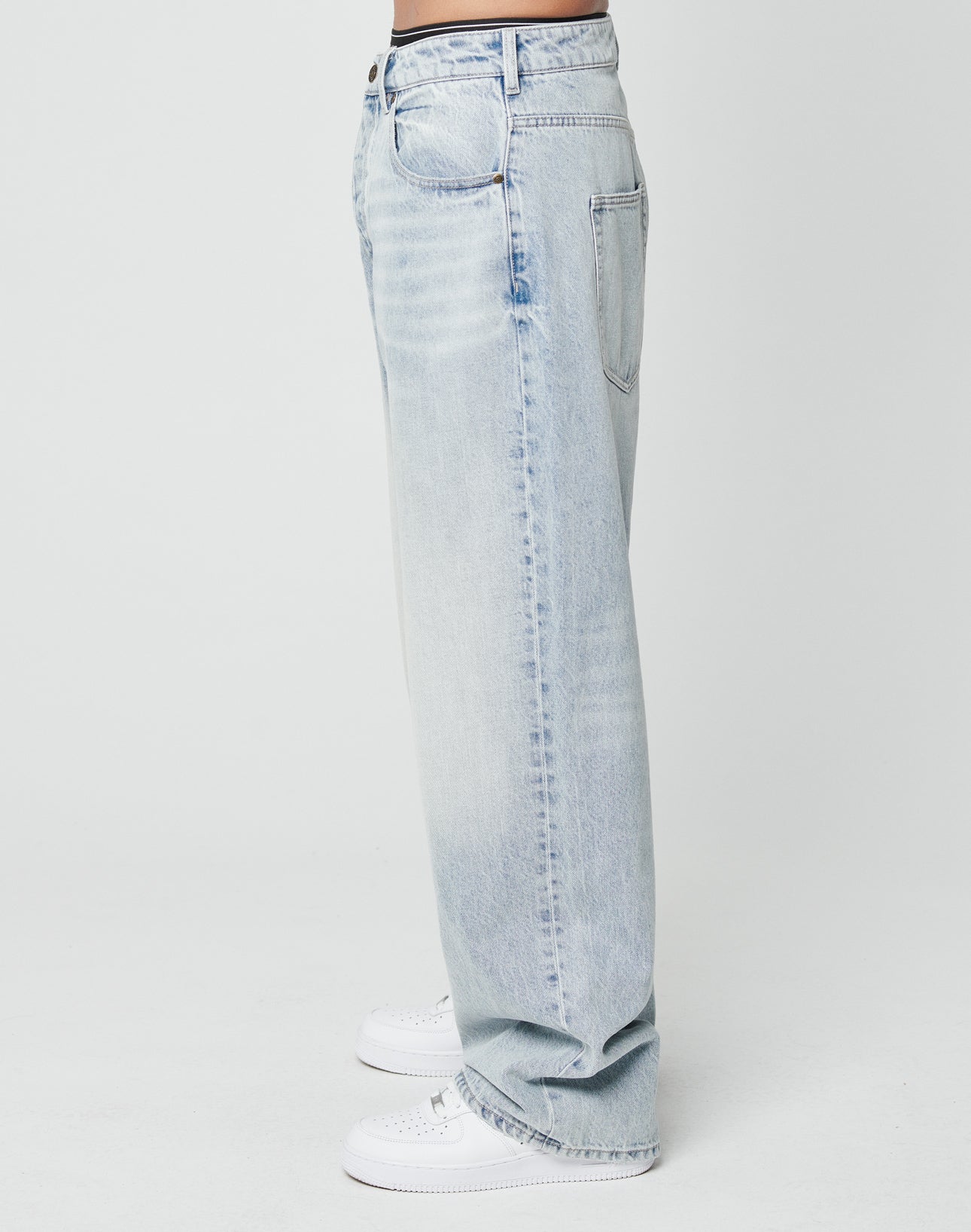 A person stands in profile wearing LFDY Baggy Denim by LIVE FAST DIE YOUNG—light blue, straight-leg 100% cotton jeans in a baggy fit—paired with white sneakers against a plain white background.