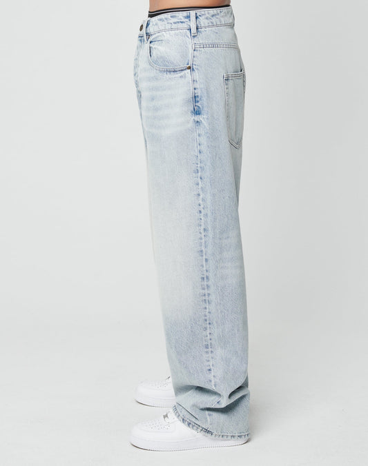 A person stands in profile wearing LFDY Baggy Denim by LIVE FAST DIE YOUNG—light blue, straight-leg 100% cotton jeans in a baggy fit—paired with white sneakers against a plain white background.