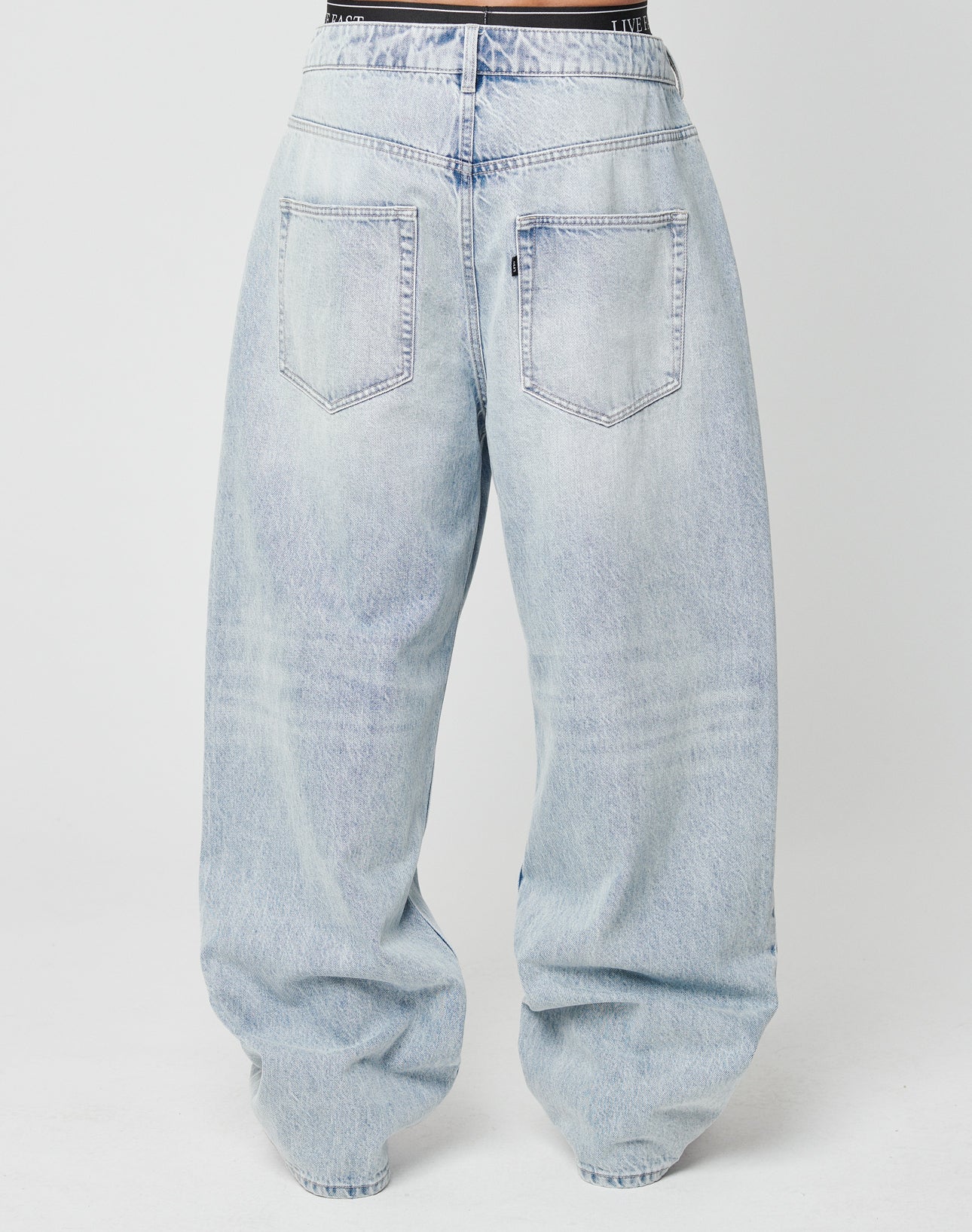 Back view of a person wearing LIVE FAST DIE YOUNG LFDY Baggy Denim—light blue, high-waisted jeans with a baggy fit, wide straight legs, two large back pockets, slight ankle crinkles, and made from 100% Baumwolle.