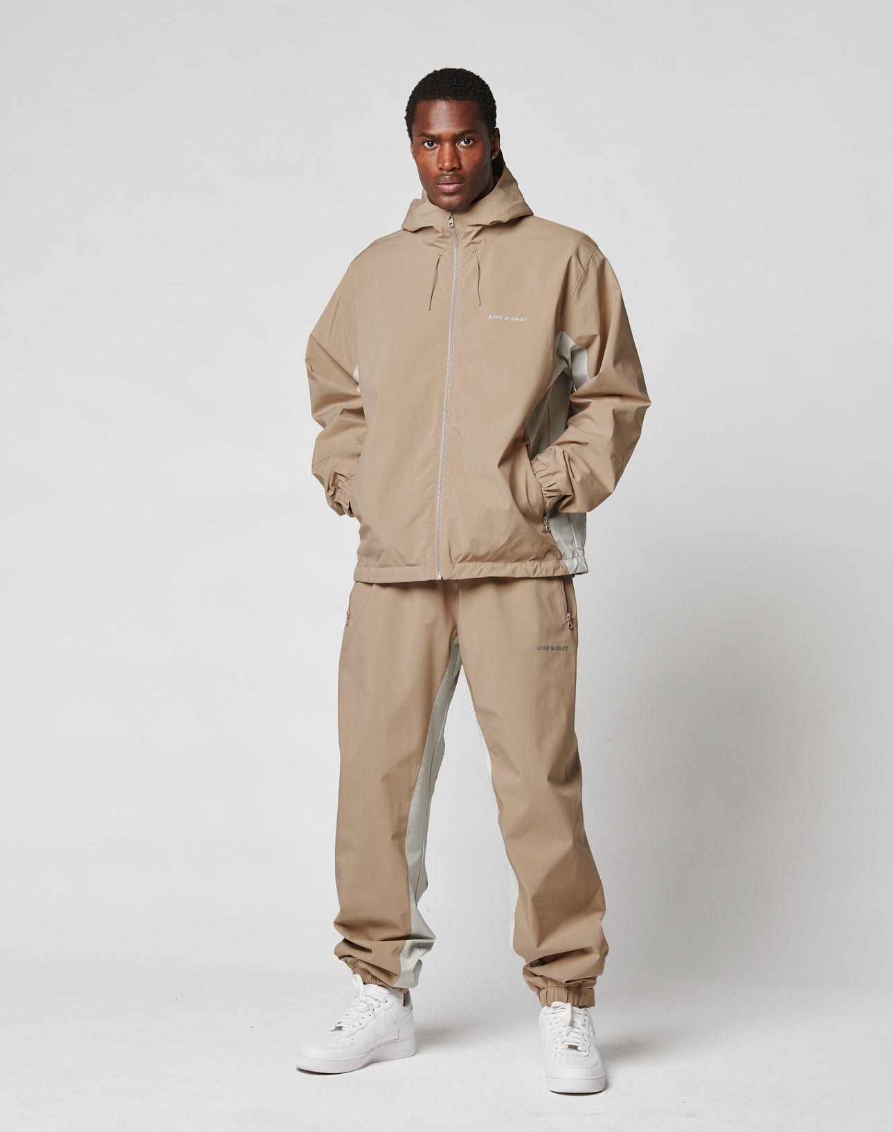 A man stands against a plain white background wearing LIVE FAST DIE YOUNG Basic Shell Pants and white sneakers, hands in pockets, facing forward with a neutral expression.