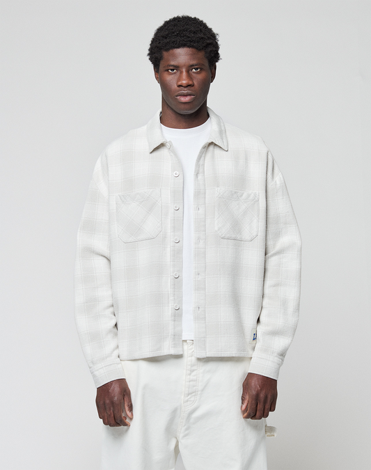 A man with short, curly hair and a neutral expression stands facing forward, wearing a LIVE FAST DIE YOUNG Vintage Flannel in an oversize fit over a white T-shirt and white pants against a plain light gray background. | model
