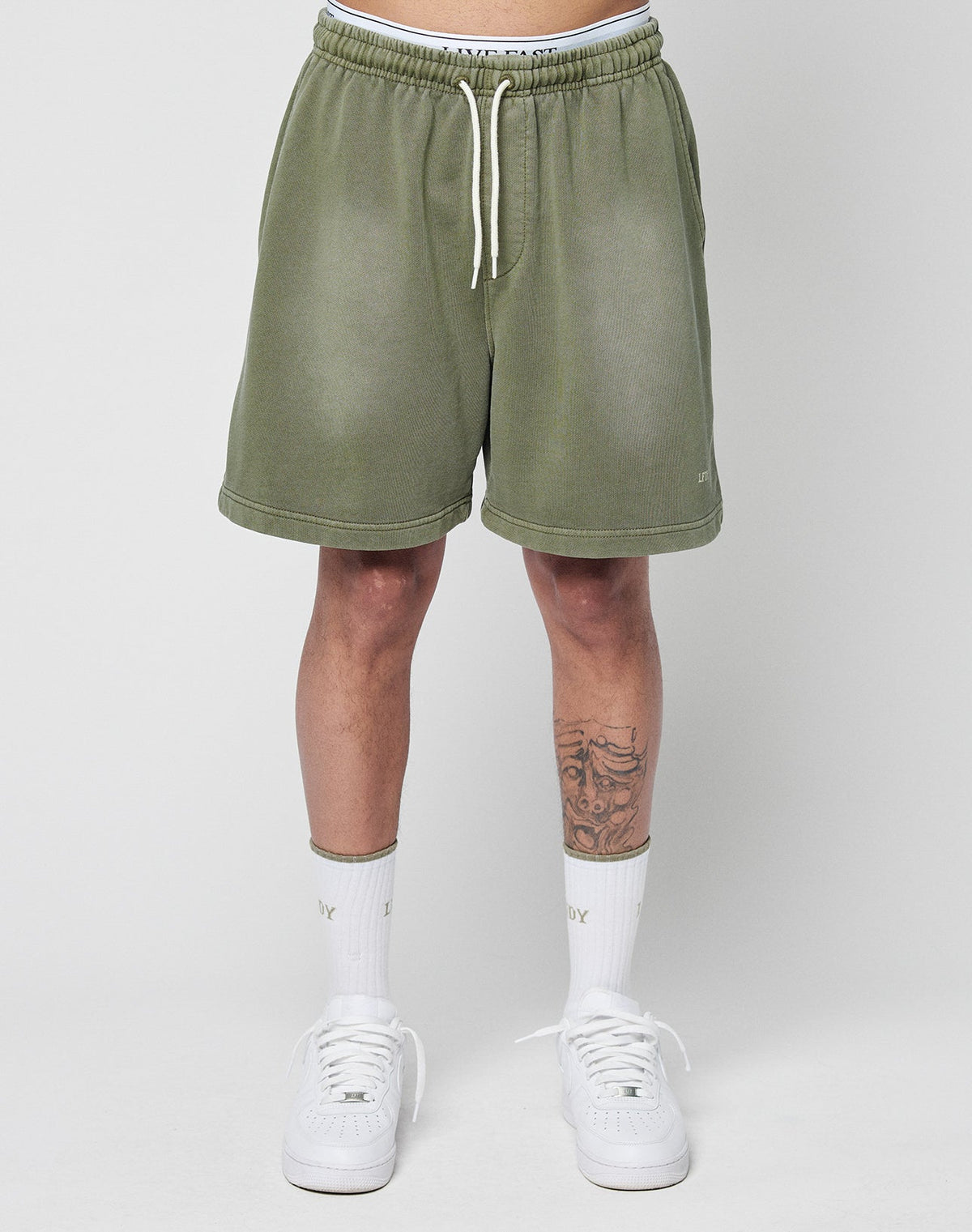 A person wears LIVE FAST DIE YOUNG Basic Sweat Shorts in olive green with an elastic waistband, white crew socks, and white sneakers. Against a plain light background, only their lower torso and legs—with a tattoo on the right shin—are visible.
