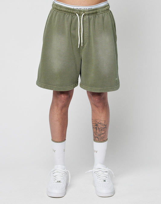 A person wears LIVE FAST DIE YOUNG Basic Sweat Shorts in olive green with an elastic waistband, white crew socks, and white sneakers. Against a plain light background, only their lower torso and legs—with a tattoo on the right shin—are visible.