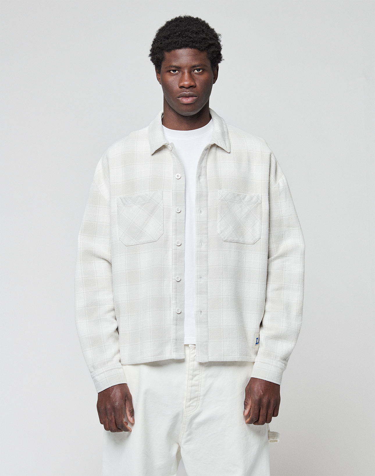 A man with short, curly hair and a neutral expression stands facing forward, wearing a LIVE FAST DIE YOUNG Vintage Flannel in an oversize fit over a white T-shirt and white pants against a plain light gray background. | model