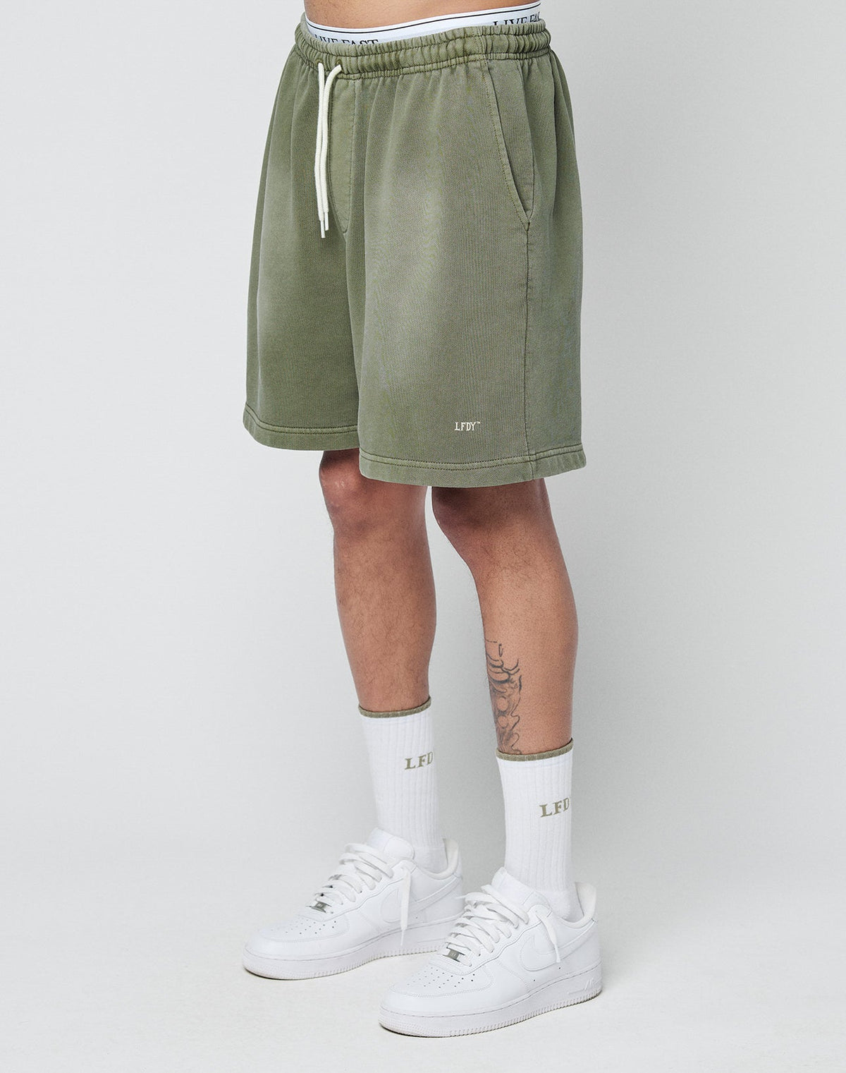 A person wears LIVE FAST DIE YOUNG Basic Sweat Shorts in olive green with an elastic waistband, paired with white LEFIT socks and white sneakers, standing against a plain light gray background. Only the lower torso and legs are visible. | model