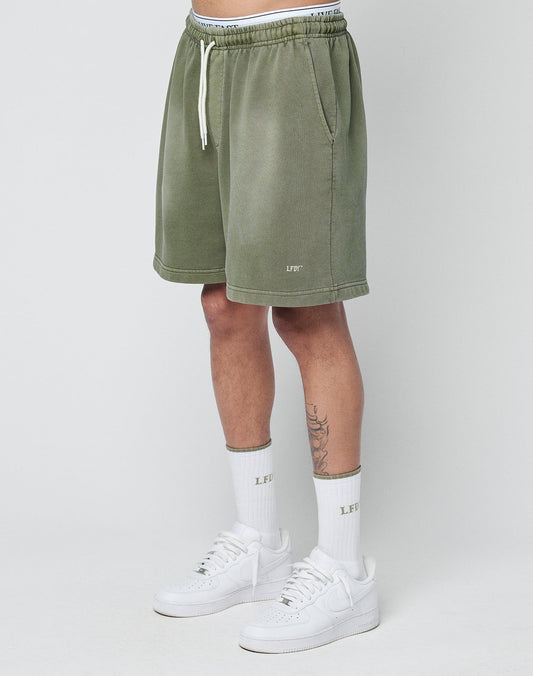 A person wears LIVE FAST DIE YOUNG Basic Sweat Shorts in olive green with an elastic waistband, paired with white LEFIT socks and white sneakers, standing against a plain light gray background. Only the lower torso and legs are visible. | model
