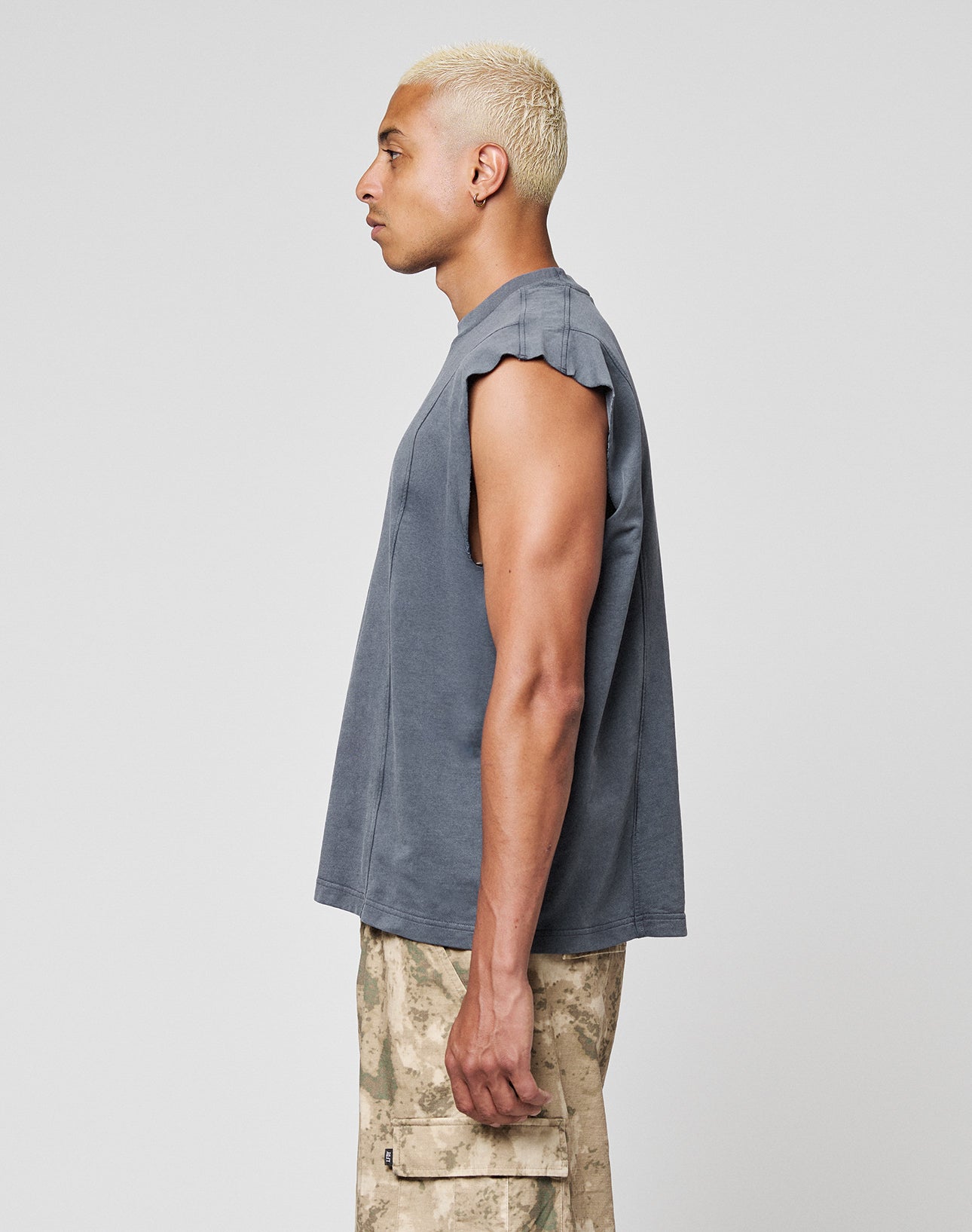 A person with short blond hair stands in profile against a plain light background wearing a LIVE FAST DIE YOUNG Heavy Muscle Tank in an oversize fit and sleeveless style, paired with beige camouflage cargo pants.