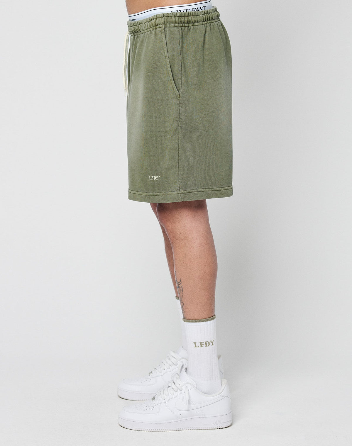 A person stands sideways, showing only their lower body, wearing LIVE FAST DIE YOUNG Basic Sweat Shorts in olive green with a regular fit, elastic waistband, pockets, a small LFDY logo near the hem, plus white sneakers and LFDY socks.