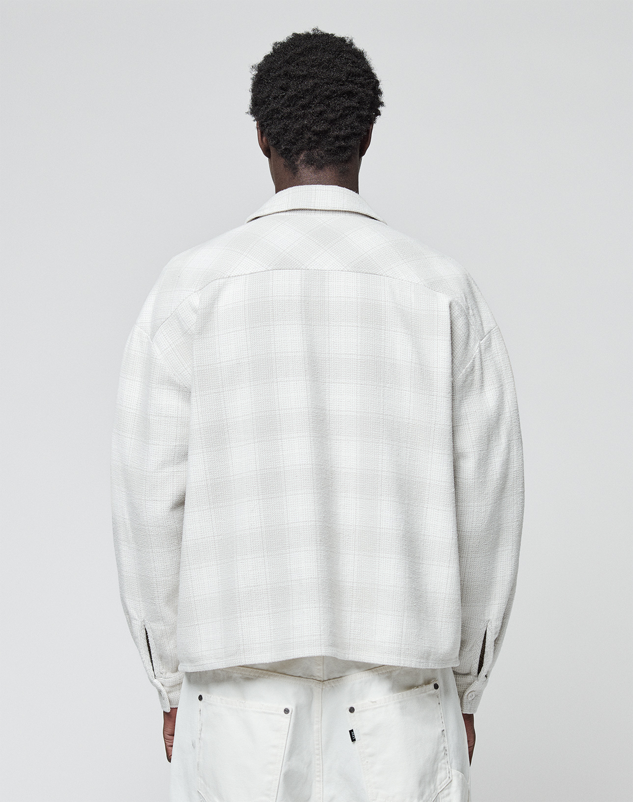 A person with short curly hair stands with their back to the camera, wearing a light plaid Vintage Flannel from LIVE FAST DIE YOUNG and white pants against a plain, light background.
