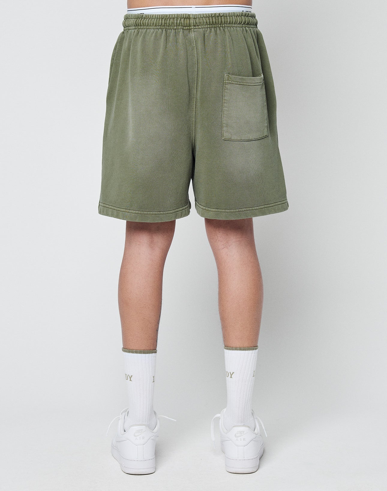A person is seen from behind wearing LIVE FAST DIE YOUNG Basic Sweat Shorts in olive green with an elastic waistband and back pocket, paired with white crew socks and sneakers, set against a plain light background.