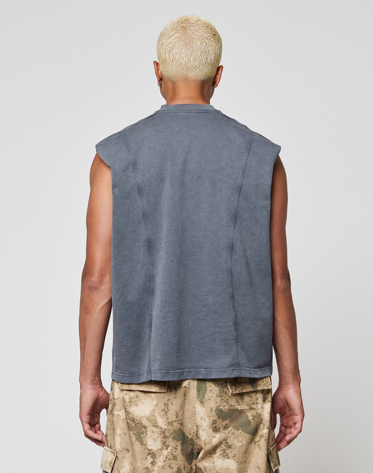 A person with short, bleached blond hair is shown from behind wearing a LIVE FAST DIE YOUNG Heavy Muscle Tank in an oversize fit and beige camouflage cargo shorts, set against a plain light gray background.