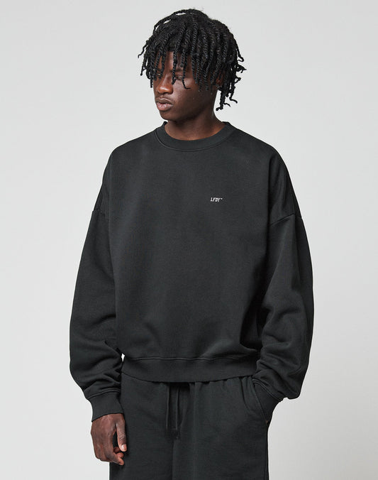 A person with medium-length, twisted hair wears the LIVE FAST DIE YOUNG Basic Evidence Sweater—an oversize black Baumwolle Polyester sweatshirt with small white chest text—paired with matching black pants against a plain light gray background.