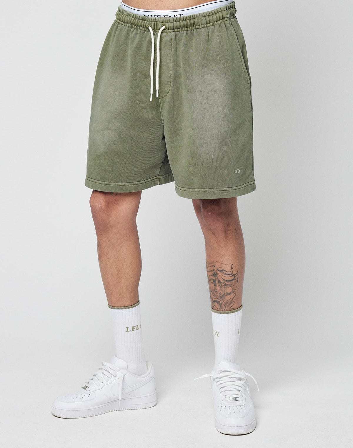 A person wears LIVE FAST DIE YOUNG Basic Sweat Shorts in olive green with an elastic waistband and white drawstring, paired with white socks and sneakers. Their lower legs and a right-shin tattoo are visible against a plain background.