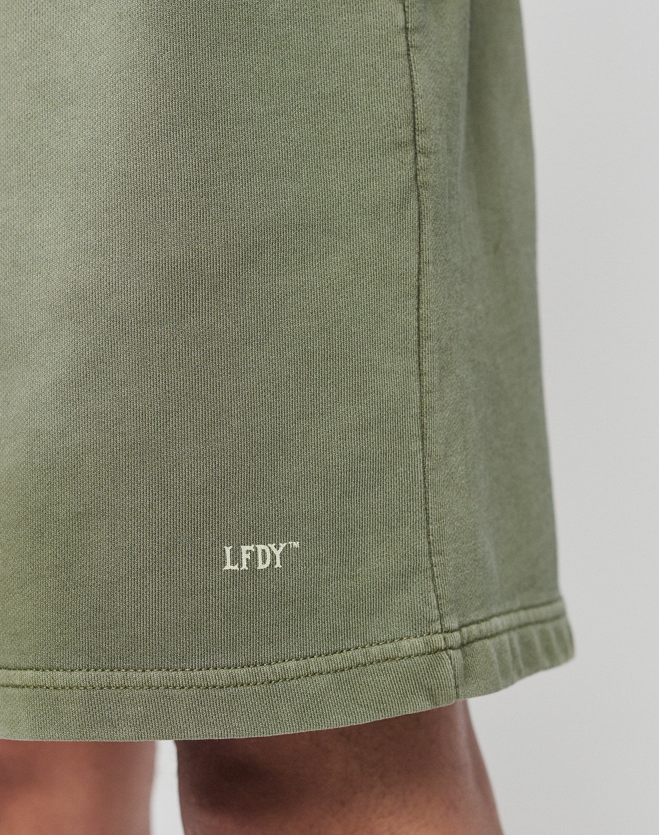 Close-up of the hem of green Basic Sweat Shorts by LIVE FAST DIE YOUNG, featuring small white LFDY text near the bottom edge. Part of a persons leg is visible below the hem.