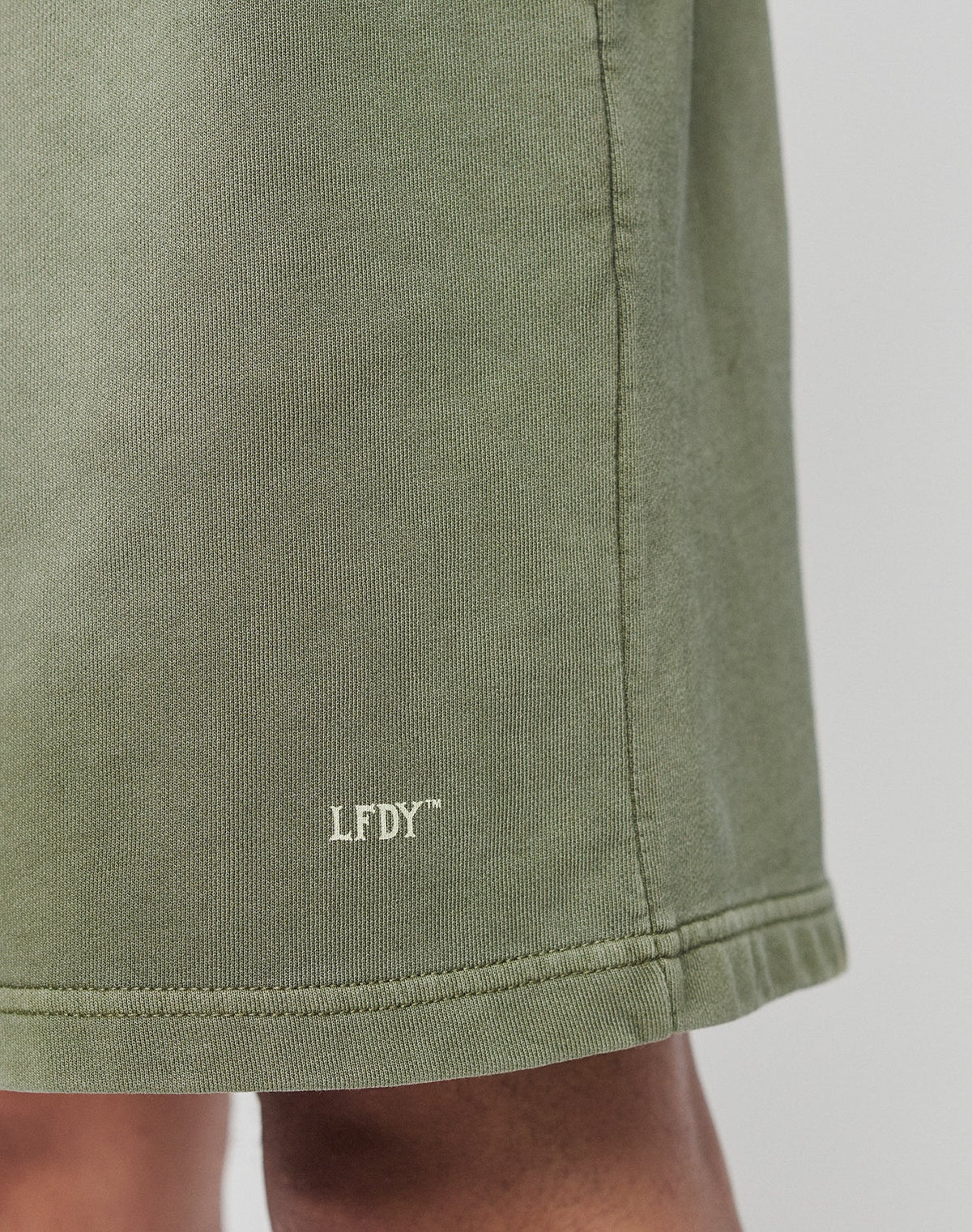 Close-up of the hem of green Basic Sweat Shorts by LIVE FAST DIE YOUNG, featuring small white LFDY text near the bottom edge. Part of a persons leg is visible below the hem.
