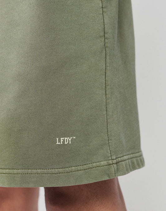Close-up of the hem of green Basic Sweat Shorts by LIVE FAST DIE YOUNG, featuring small white LFDY text near the bottom edge. Part of a persons leg is visible below the hem.