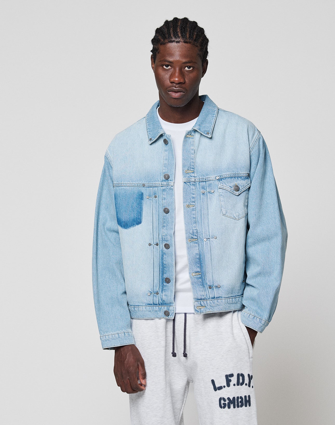 A man wears a LIGHT FAST DIE YOUNG LFDY Trucker Jacket in light blue denim over a white t-shirt and light gray Regular Fit Baumwolle sweatpants with text on the left leg, standing against a plain light gray background.
