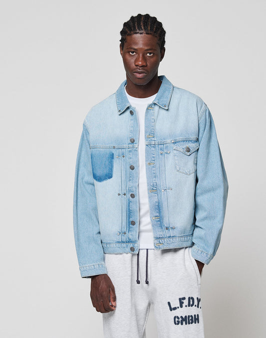 A man wears a LIGHT FAST DIE YOUNG LFDY Trucker Jacket in light blue denim over a white t-shirt and light gray Regular Fit Baumwolle sweatpants with text on the left leg, standing against a plain light gray background.
