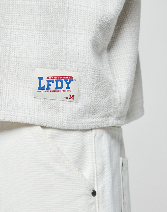 A close-up of a person wearing a textured off-white LIVE FAST DIE YOUNG Vintage Flannel with an oversize fit and white pants. The label reads LFDY OFFICIALLY LICENSED PRODUCT with a red M for the size.