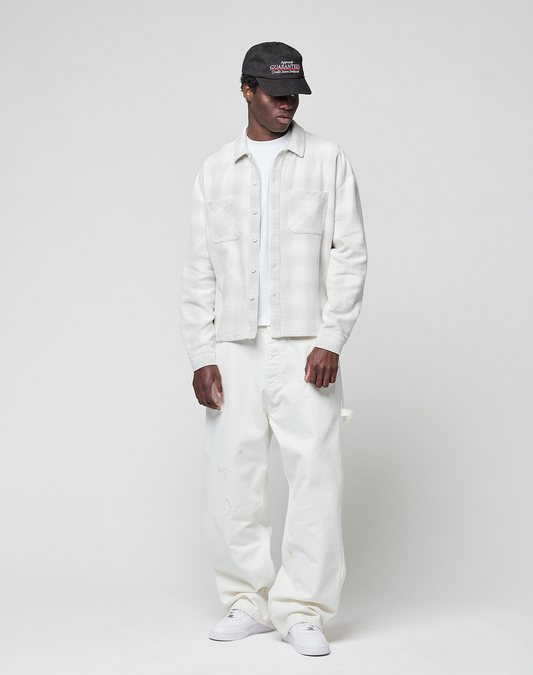 A person in a black cap, LIVE FAST DIE YOUNG Vintage Flannel with oversize fit, loose white pants, and white sneakers stands against a plain light-gray background.