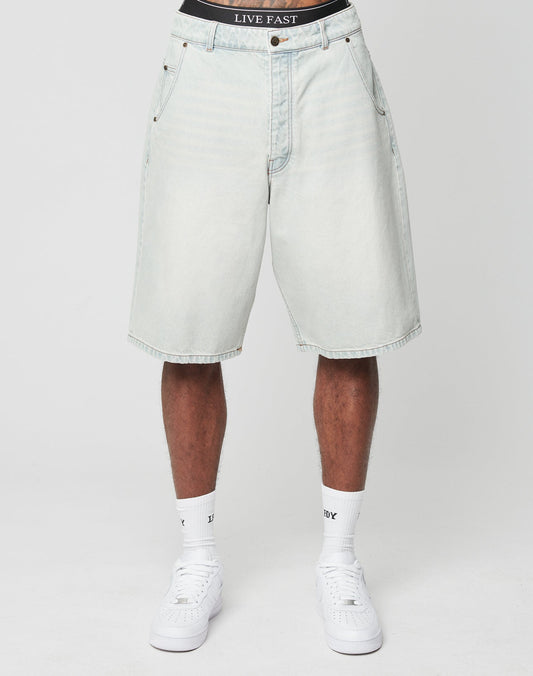 A person wearing LIVE FAST DIE YOUNG LFDY Jorts in light-wash denim with baggy fit and visible Gesäßtasche, paired with white socks, white sneakers, and black underwear featuring a LIVE FAST waistband, stands against a plain white background.