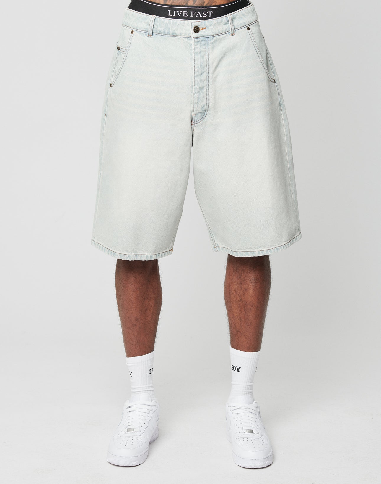 A person wearing LIVE FAST DIE YOUNG LFDY Jorts in light-wash denim with baggy fit and visible Gesäßtasche, paired with white socks, white sneakers, and black underwear featuring a LIVE FAST waistband, stands against a plain white background.